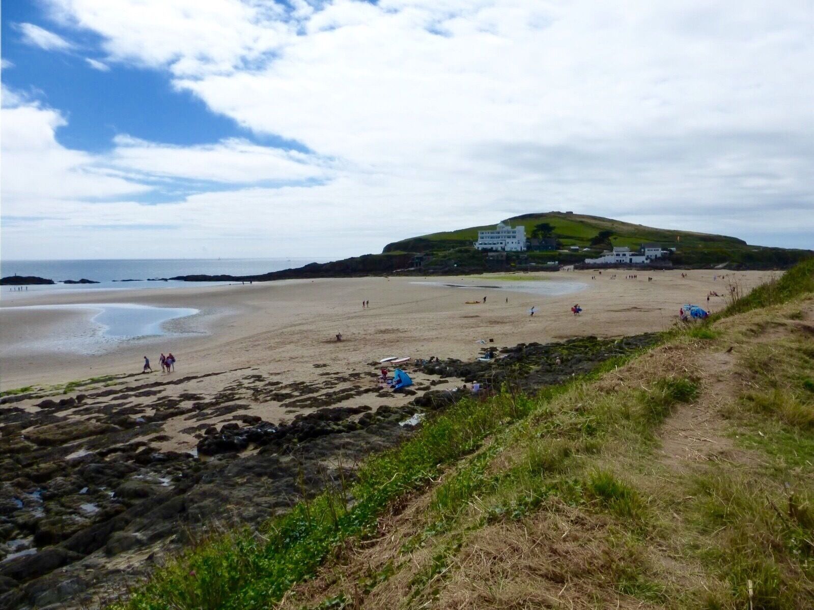 A beach with rocky outcrops, a grassy path, and a hill with buildings in the background.
