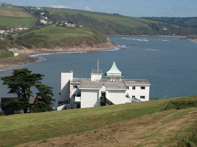 A coastal building with a green roof, overlooking a bay with a small town in the background.