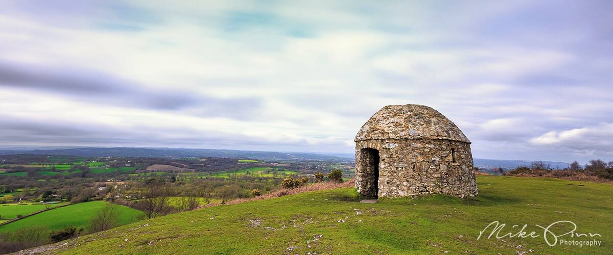 A stone structure on a grassy hill with a view of a valley and distant hills.