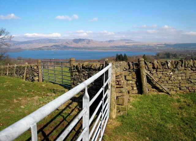 A stone wall, a metal gate, and a view of a lake with mountains in the distance.
