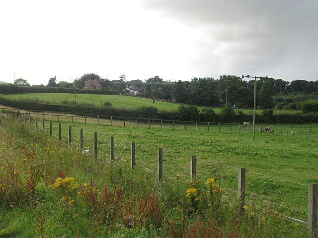A rural landscape with a fenced field, a few houses in the background, and a cloudy sky.