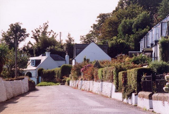 Una calle residencial tranquila bordeada de casas, una valla blanca y abundante vegetación.