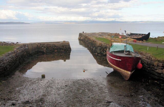 A red boat tied to a stone pier by a calm body of water, with a cloudy sky above.