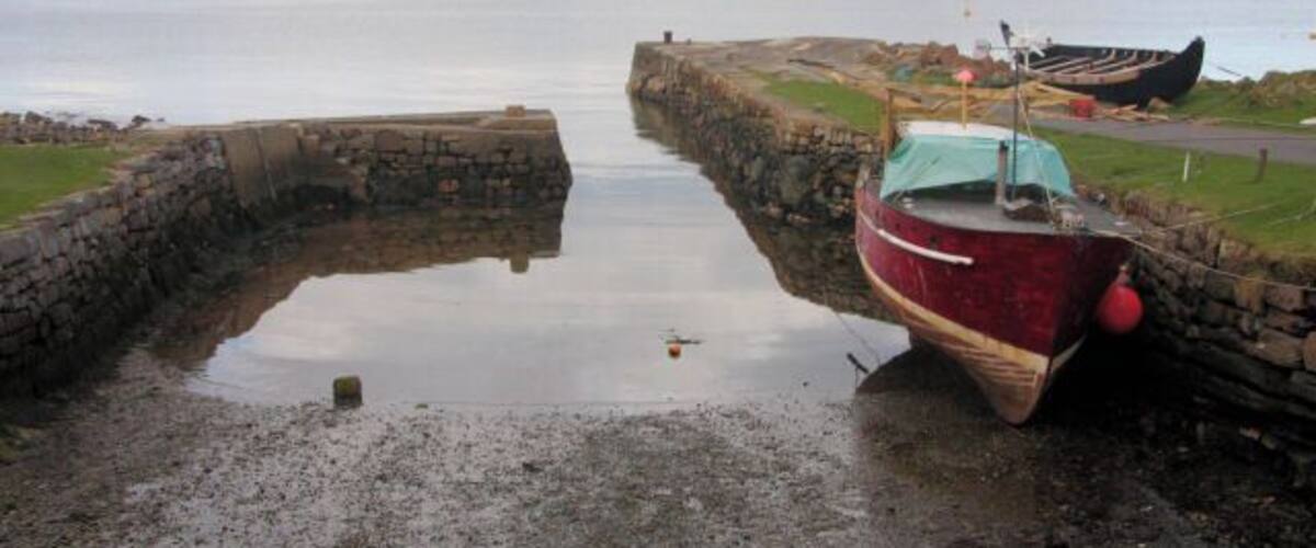 A red boat tied to a stone pier by a calm body of water, with a cloudy sky above.