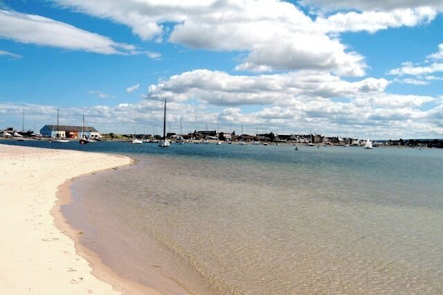A beach with clear water, a sandy shore, and a distant town with buildings and boats.