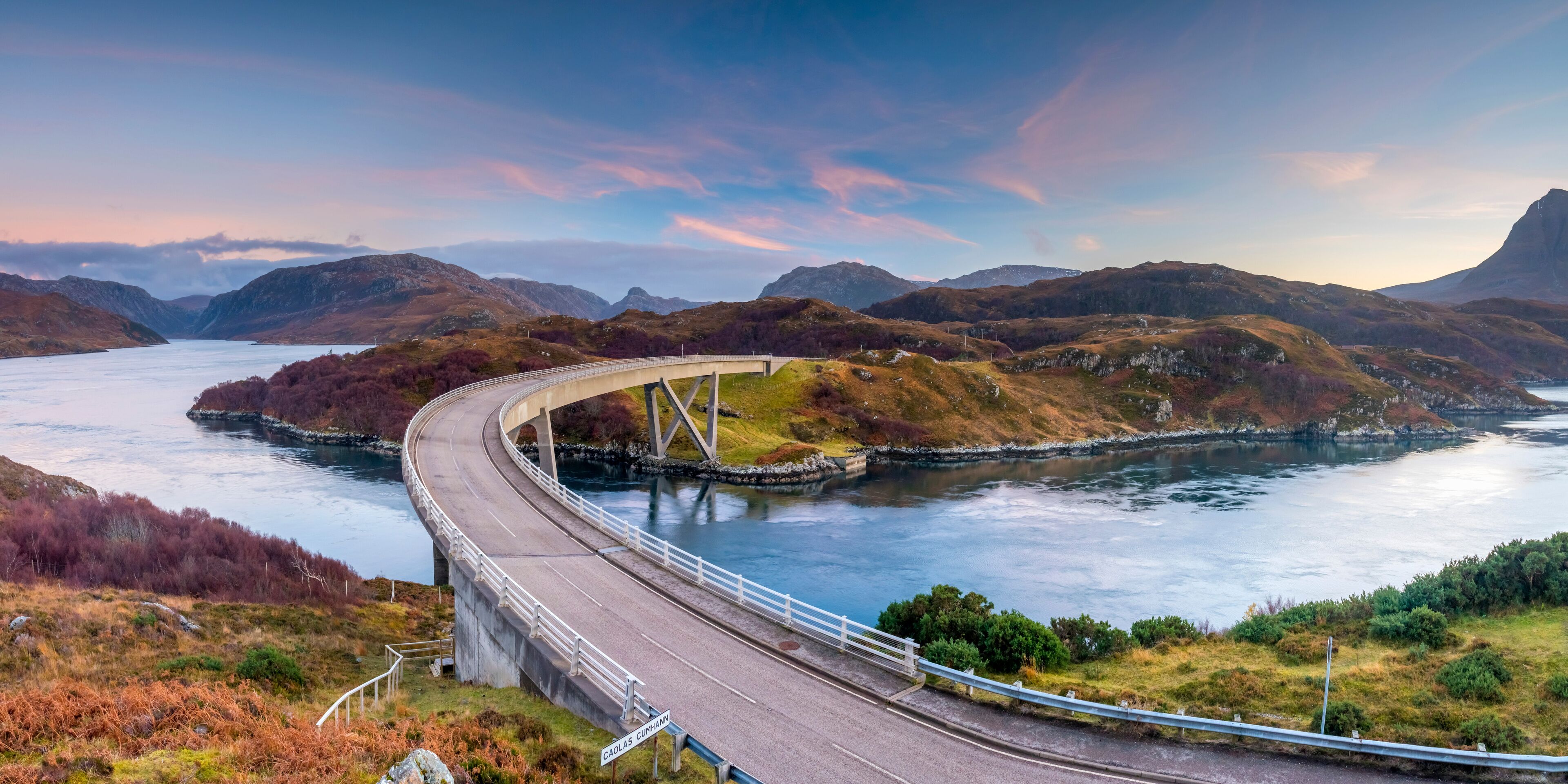 A winding road over a river, surrounded by hills and mountains.
