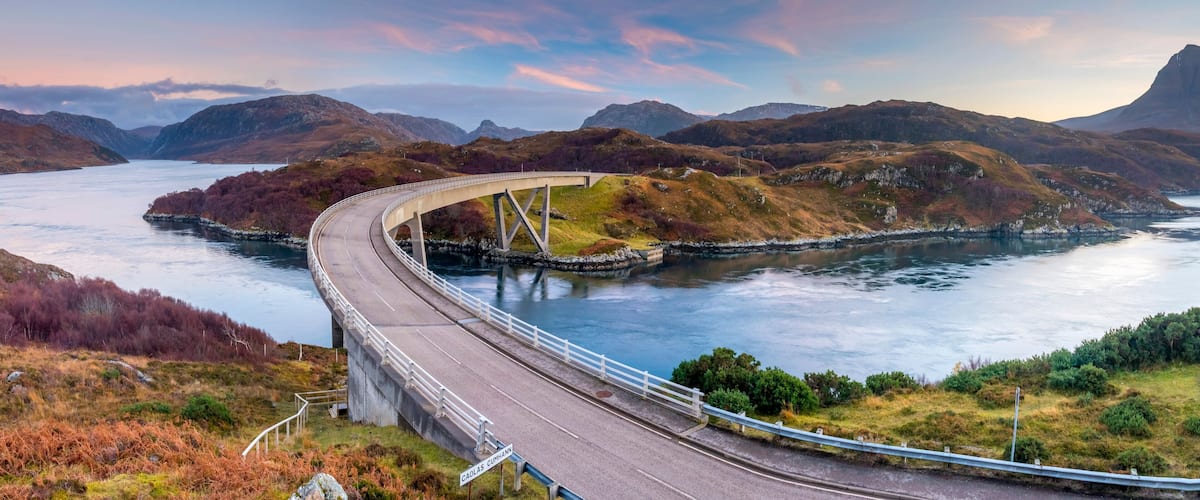 A winding road over a river, surrounded by hills and mountains.