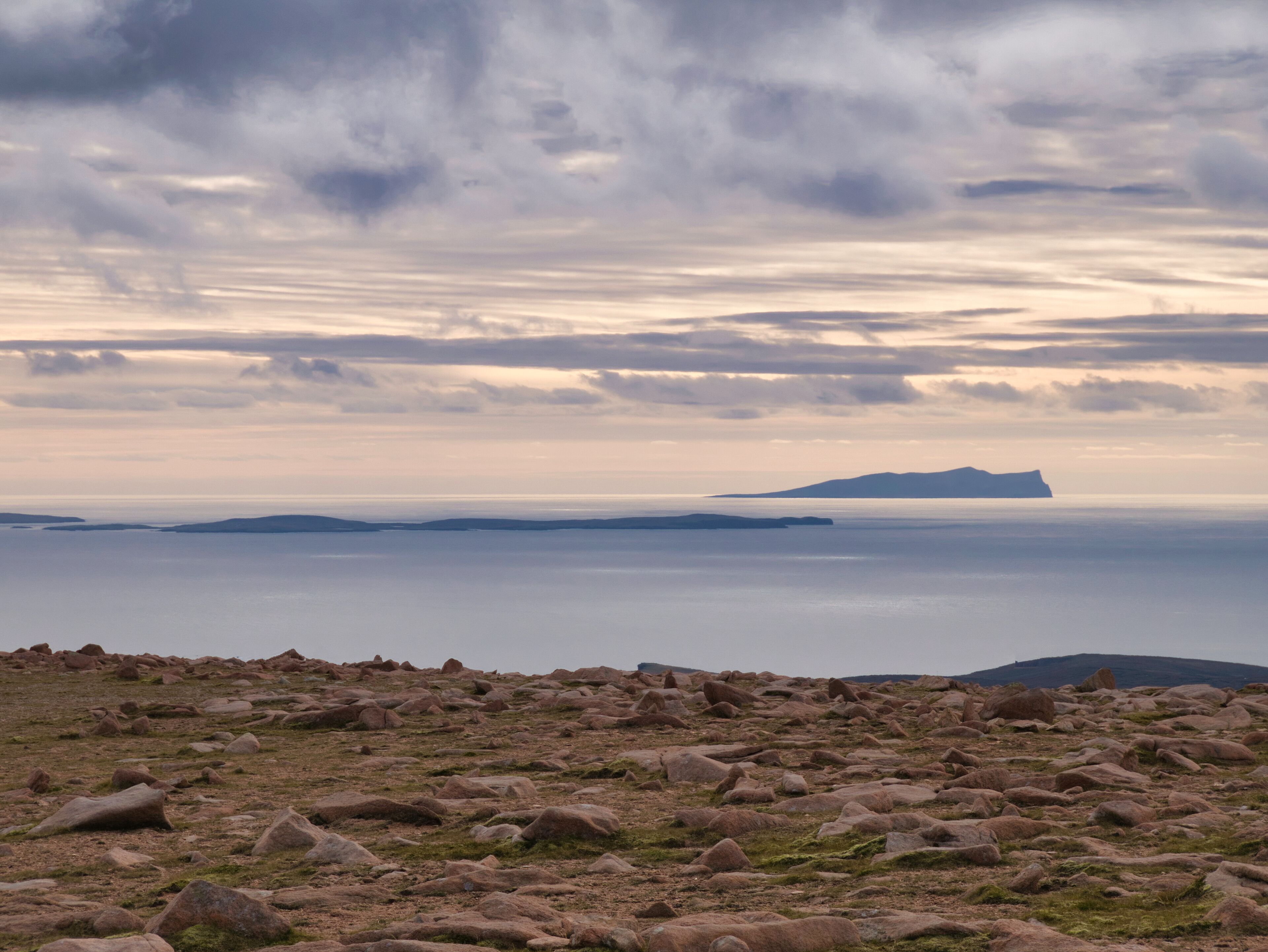 A rocky landscape with a distant island and a calm sea under a cloudy sky.