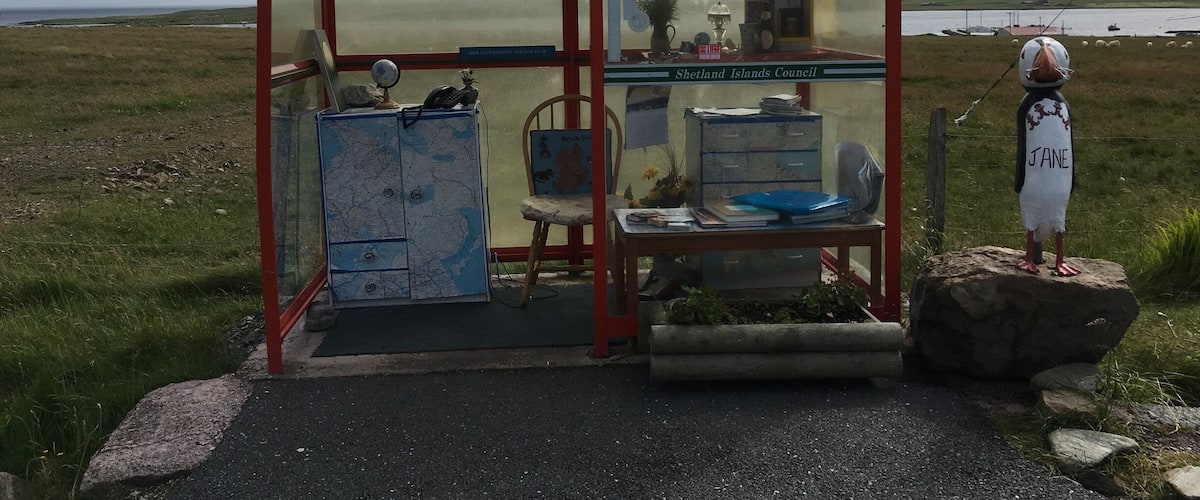 A small, rustic kiosk with a map and a chair, surrounded by a grassy landscape and a body of water in the background.
