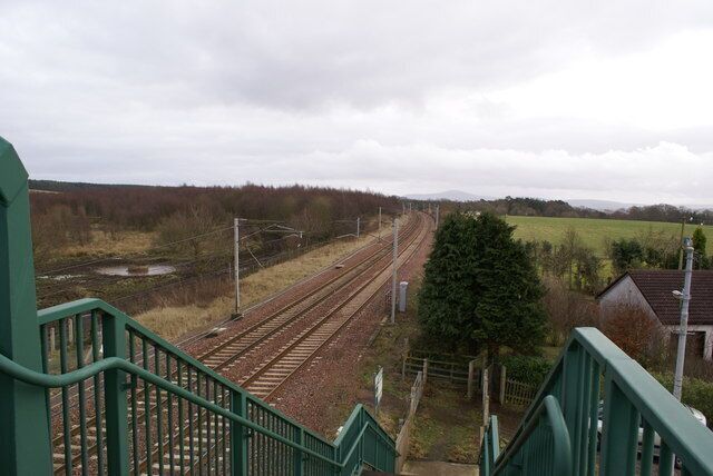 A railway station with multiple tracks, green railings, and a rural landscape with fields and trees.