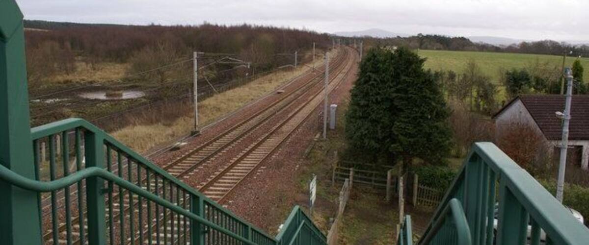 A railway station with multiple tracks, green railings, and a rural landscape with fields and trees.