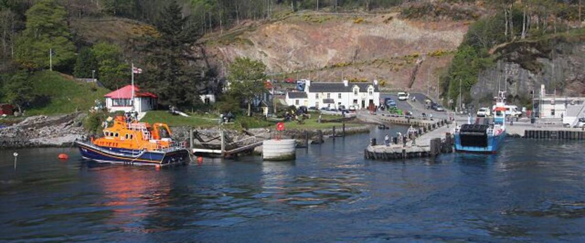 A harbor with boats, a red-roofed building, and a white building with a hill in the background.