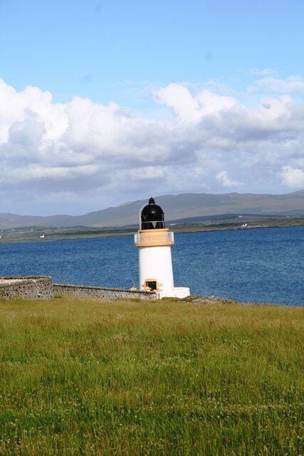 A lighthouse by a body of water with a grassy foreground and distant mountains.