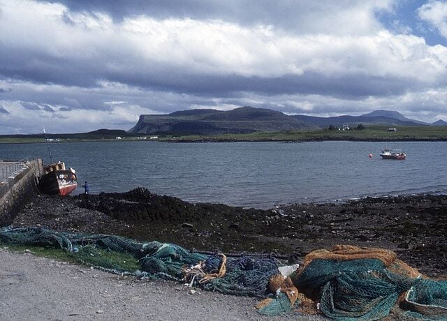 A rocky shoreline with fishing nets and a boat, a calm sea, and distant mountains under a cloudy sky.