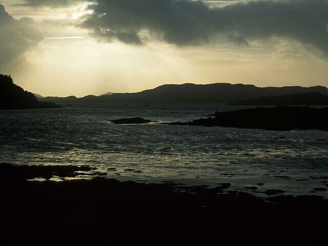 A coastal landscape with a rocky shoreline, calm sea, and distant mountains under a cloudy sky.