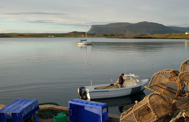 A calm lake with a small boat docked near the shore, surrounded by mountains and a distant sailboat.