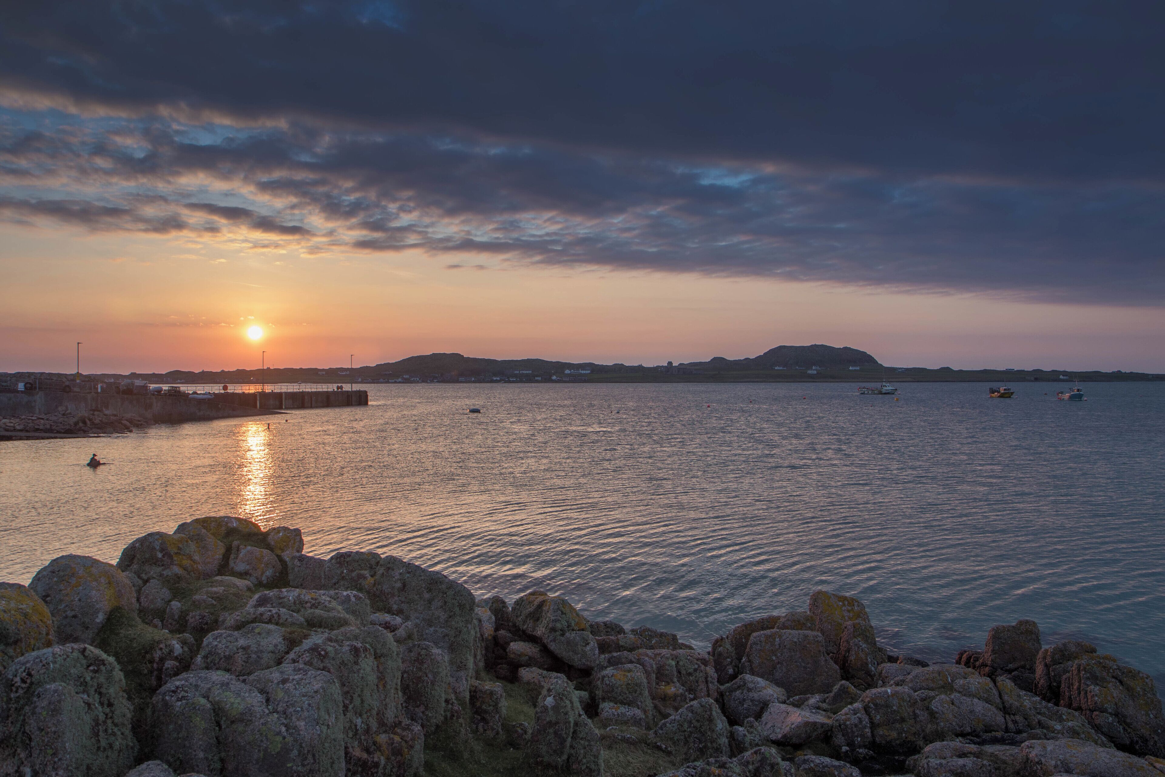 A sunset over a calm sea with rocky outcrops in the foreground and a distant shoreline.