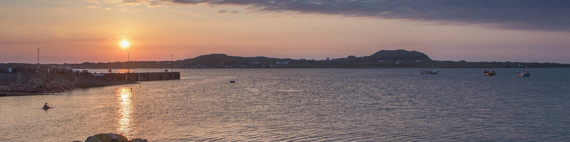 A sunset over a calm sea with rocky outcrops in the foreground and a distant shoreline.