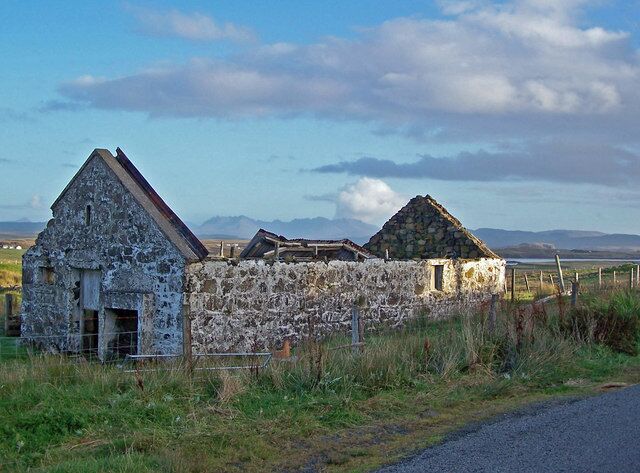 A stone wall with two stone buildings, a grassy field, and mountains in the background.