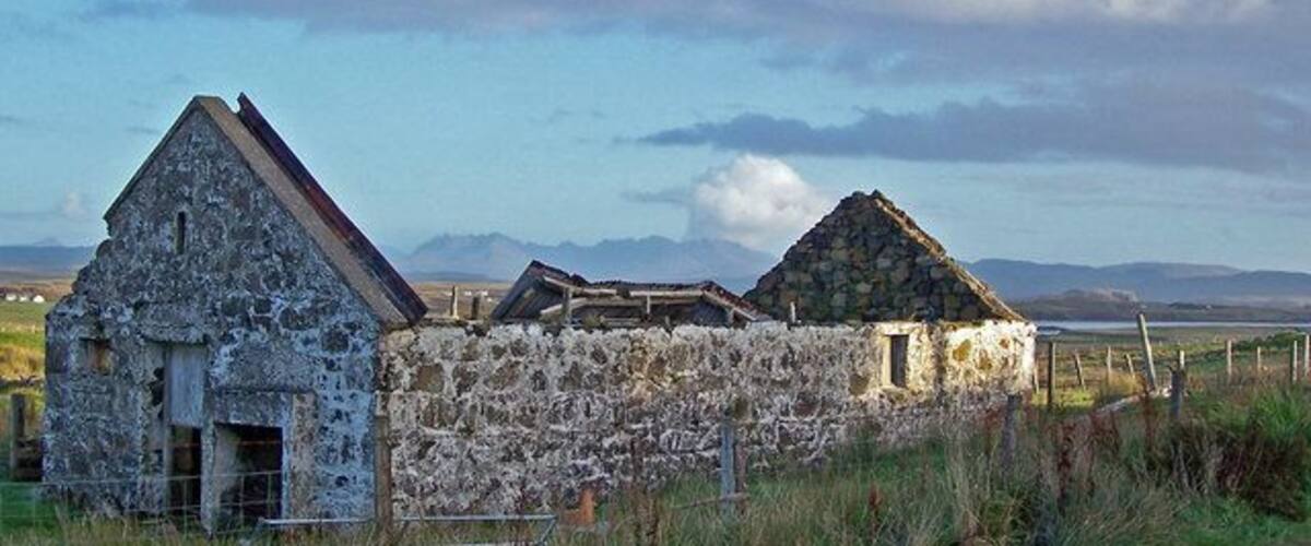 A stone wall with two stone buildings, a grassy field, and mountains in the background.