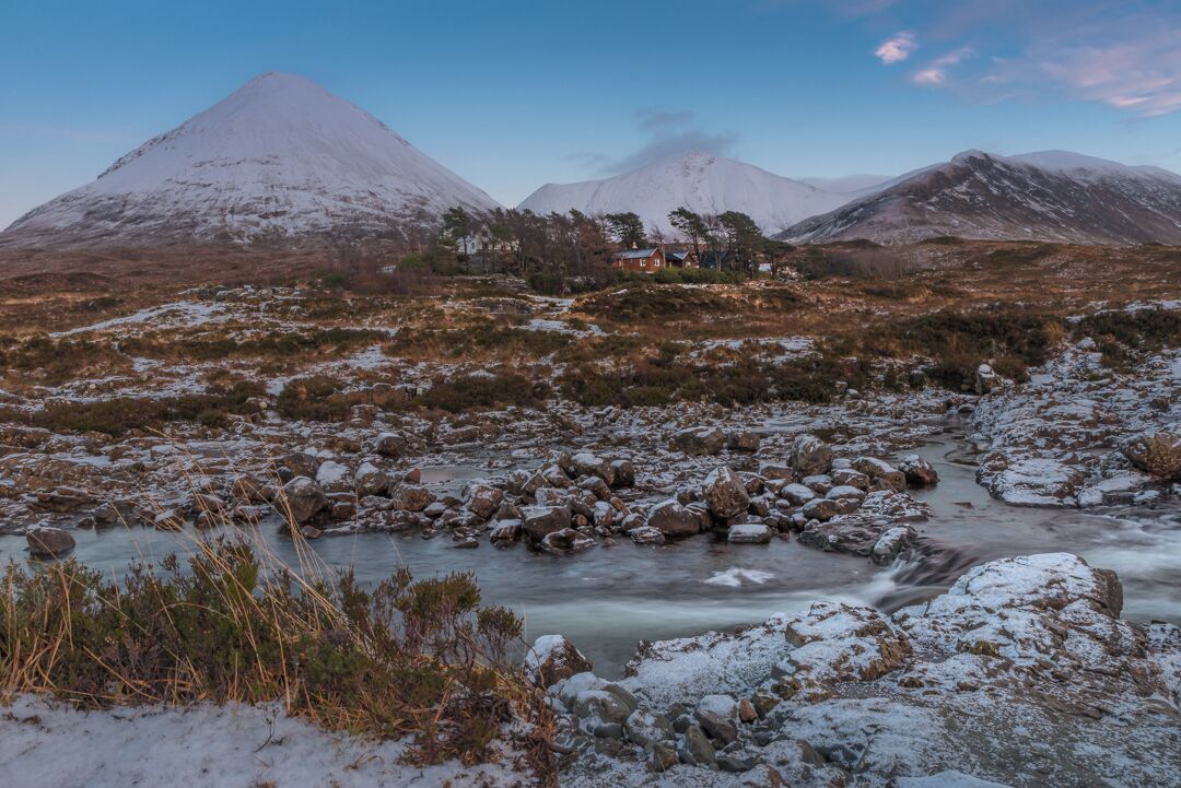 A snow-covered landscape with a river, rocky terrain, and a mountain in the background.