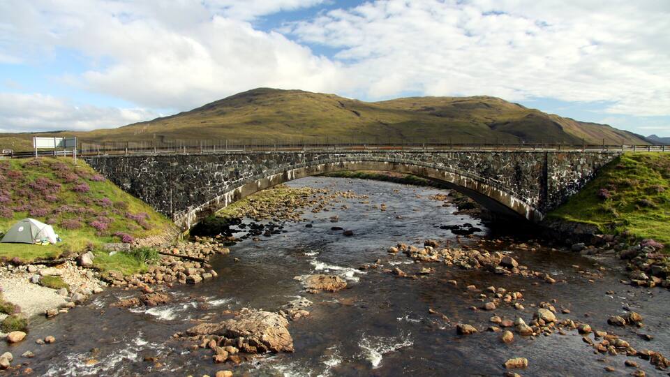 A stone bridge over a river with rocks, a tent, and hills in the background.