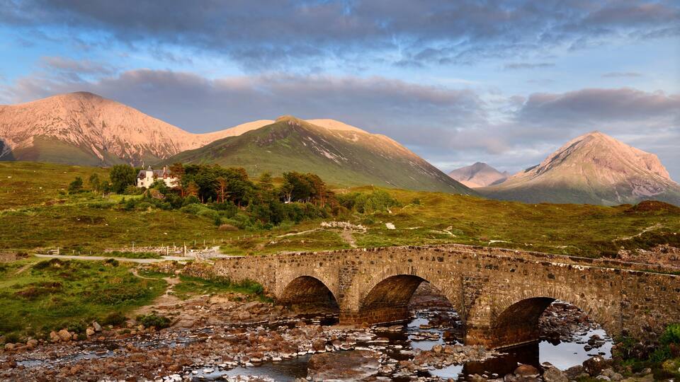 A stone bridge over a rocky stream with mountains in the background.