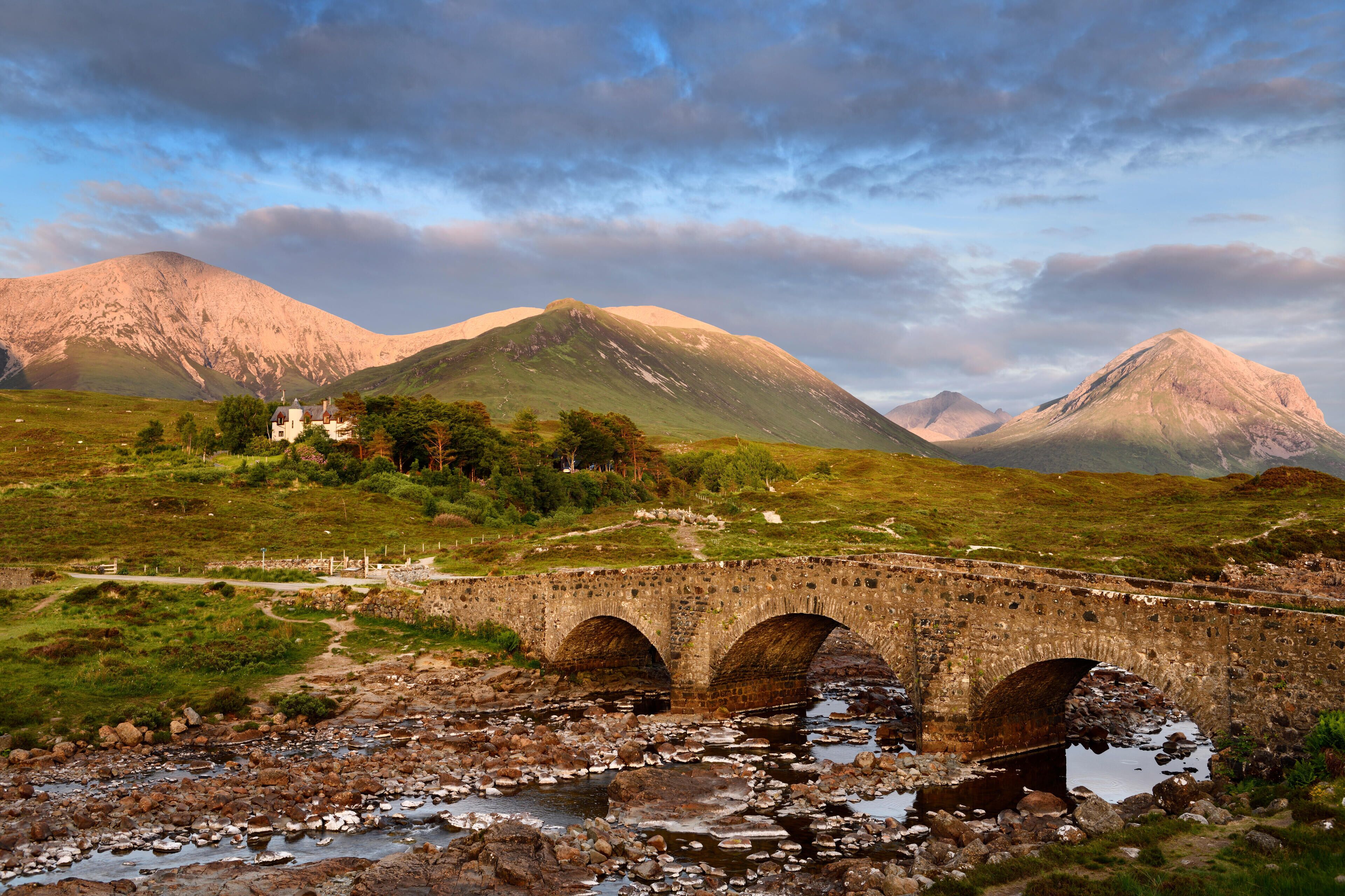 A stone bridge over a rocky stream with mountains in the background.