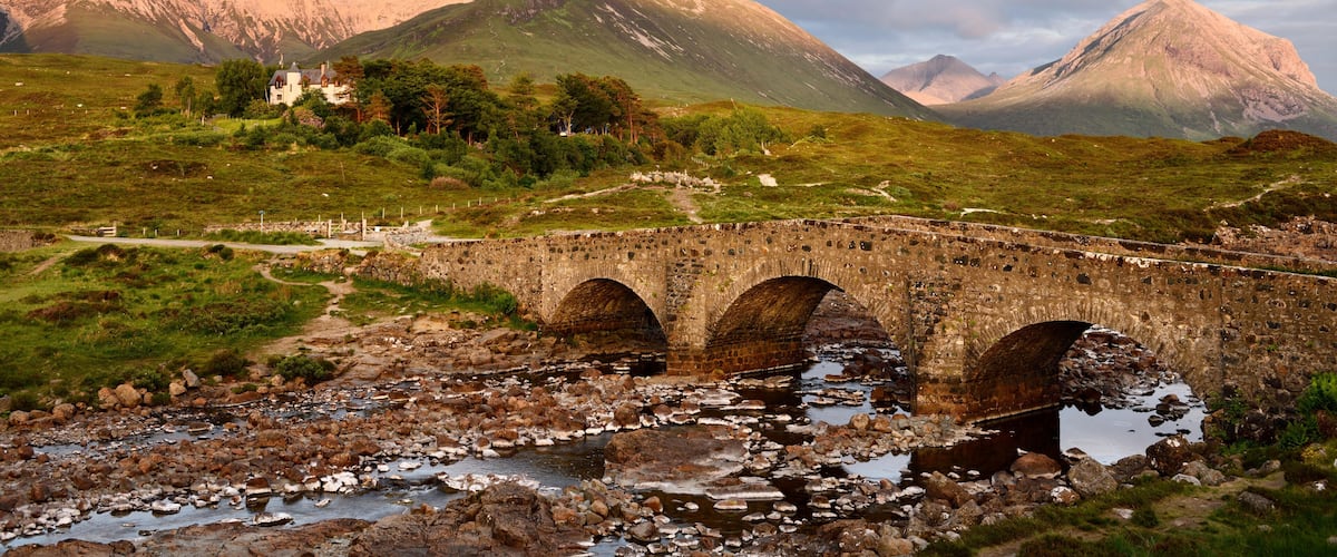 A stone bridge over a rocky stream with mountains in the background.