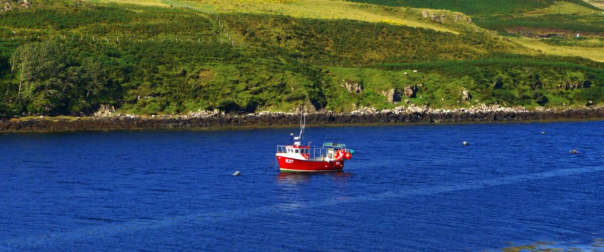 Un bote rojo sobre un mar azul tranquilo con colinas verdes al fondo.