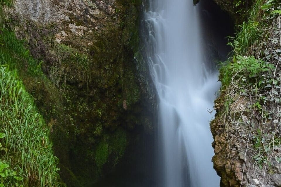 Una cascada que flueix per una gorja rocosa, amb vegetació verda i abundant als laterals.