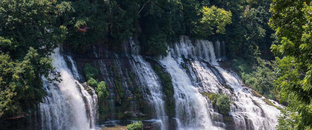 A multi-tiered waterfall cascading into a river surrounded by dense forest.