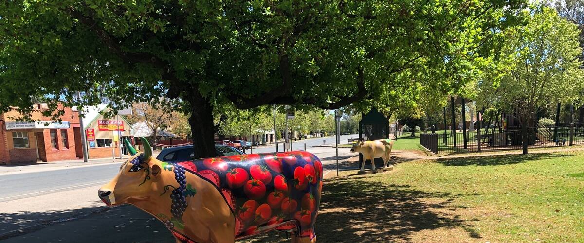 A colorful cow statue on a concrete base in a park with trees and a road in the background.