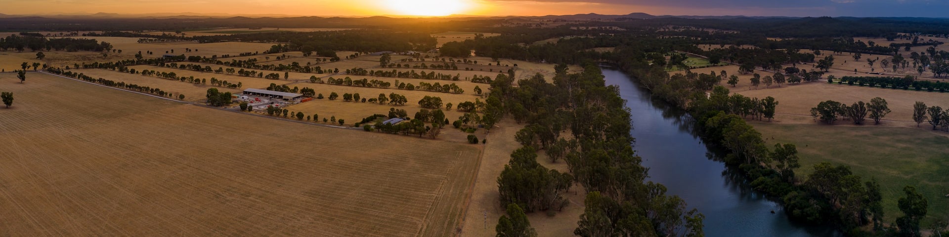 A sunset over a rural landscape with fields, a river, and scattered trees.