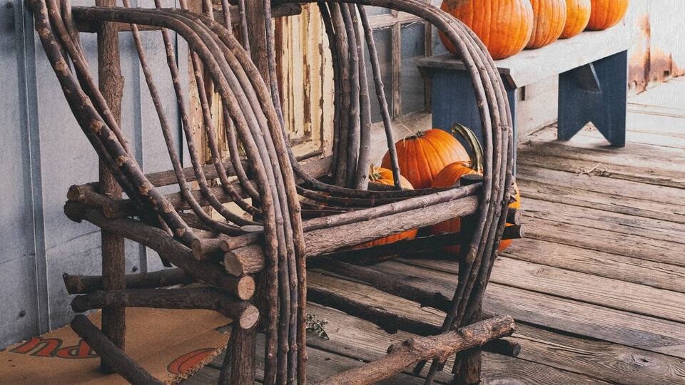 A rustic wooden chair with a woven backrest, placed on a wooden deck with a bench and pumpkins in the background.