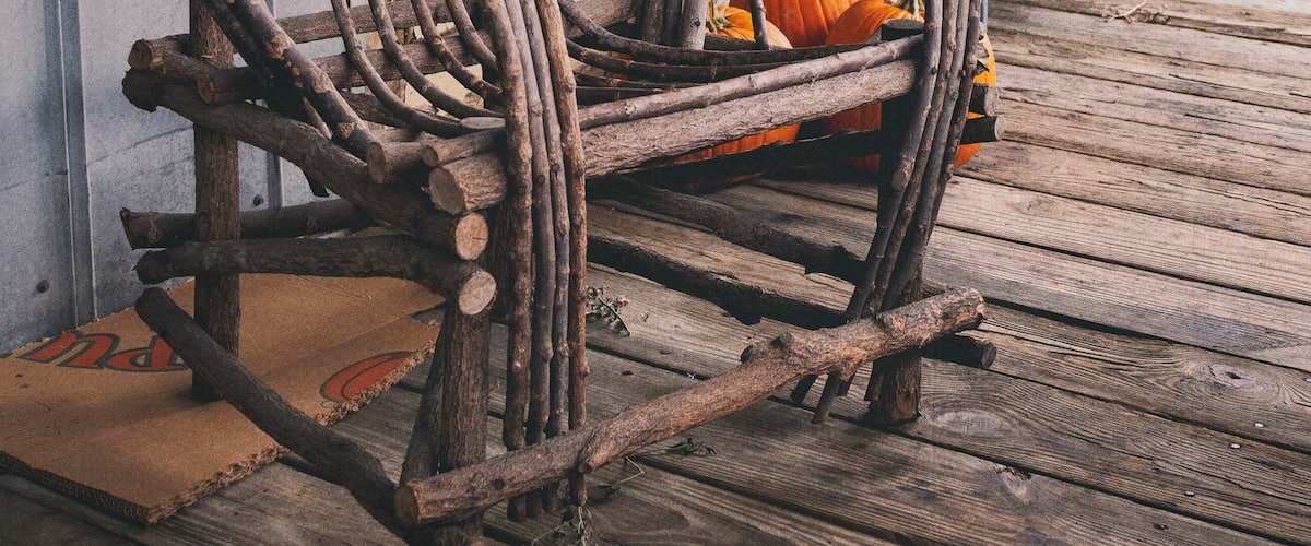 A rustic wooden chair with a woven backrest, placed on a wooden deck with a bench and pumpkins in the background.