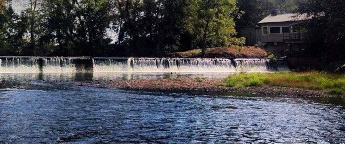 A river with a waterfall, surrounded by trees and a house in the background.