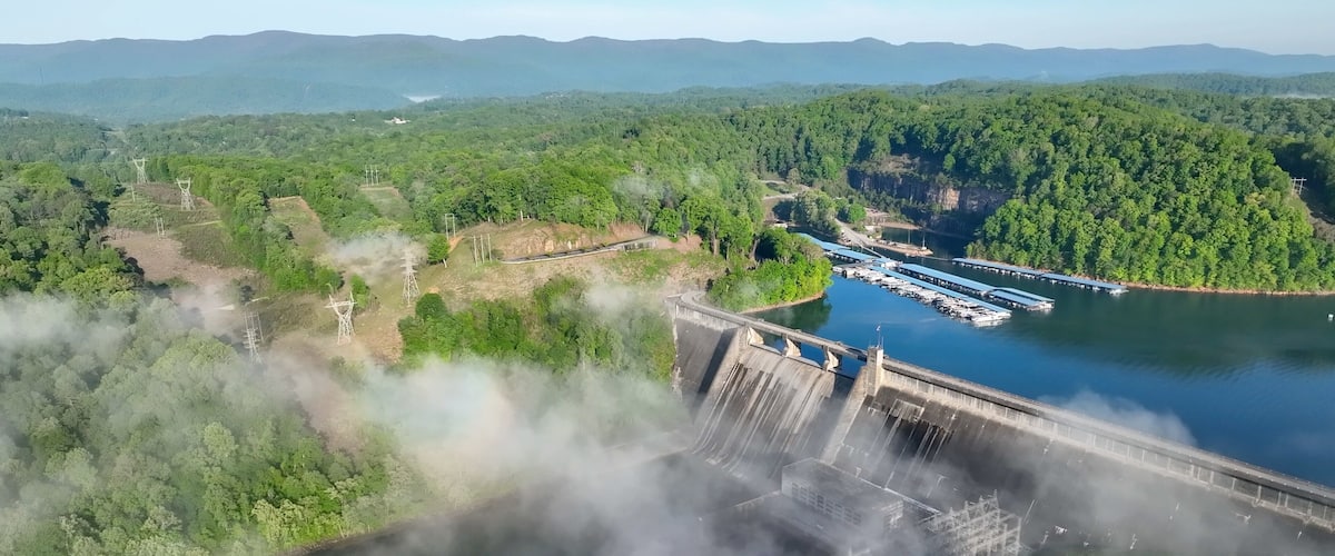 A dam with water flowing over it, surrounded by dense forest and mountains in the background.