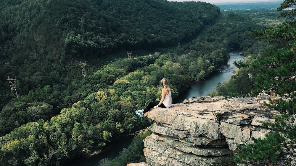 Une personne assise sur un promontoire rocheux, d’où elle a une vue sur une rivière et une vallée boisée.