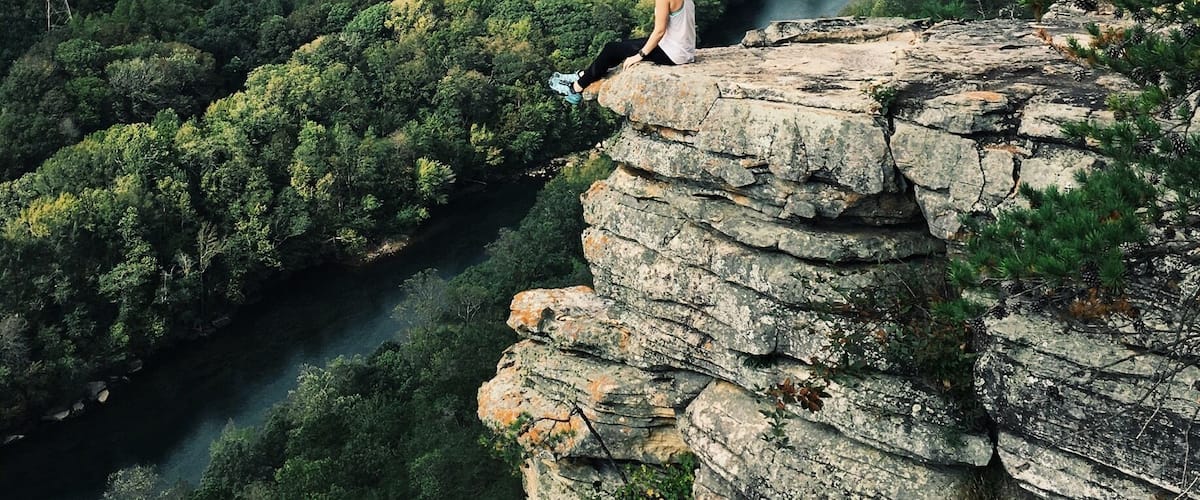 A person sitting on a rocky outcrop overlooking a river and forested valley.