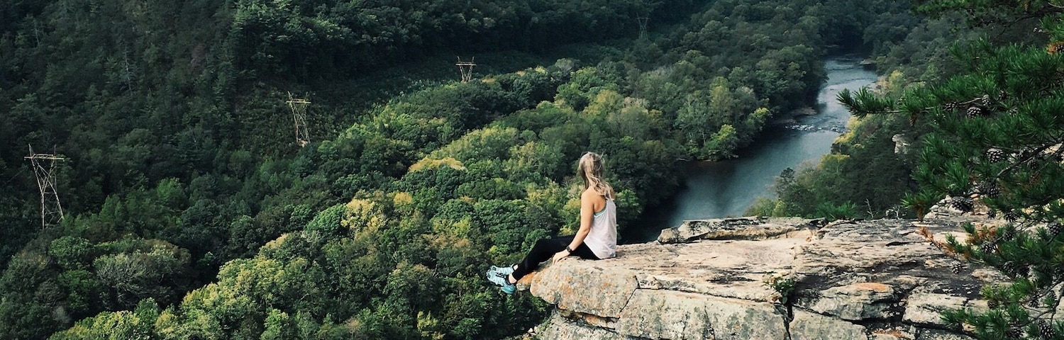 Una persona sentada sobre un afloramiento rocoso, con vista a un río y un valle cubierto de bosque.