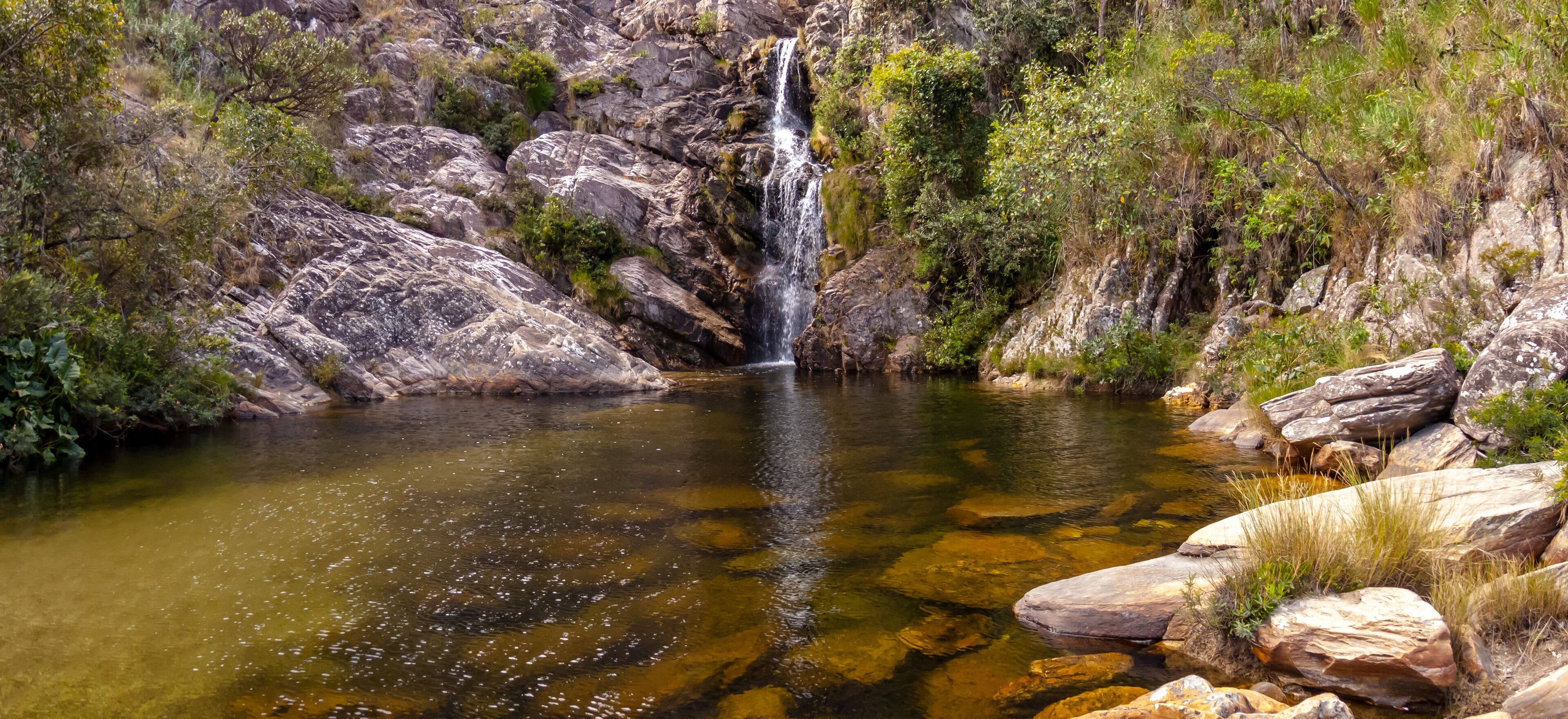 A waterfall cascading into a clear pool surrounded by rocky terrain and greenery.
