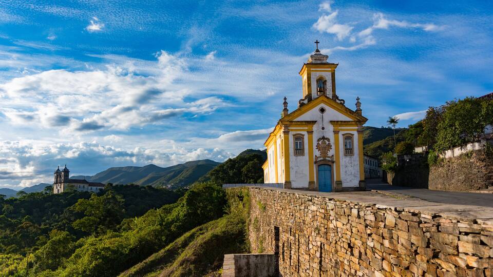 A church with a bell tower, a stone wall, and a mountain in the background.