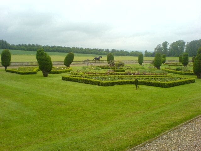 A well-manicured garden with geometric hedges and a horse in the background.
