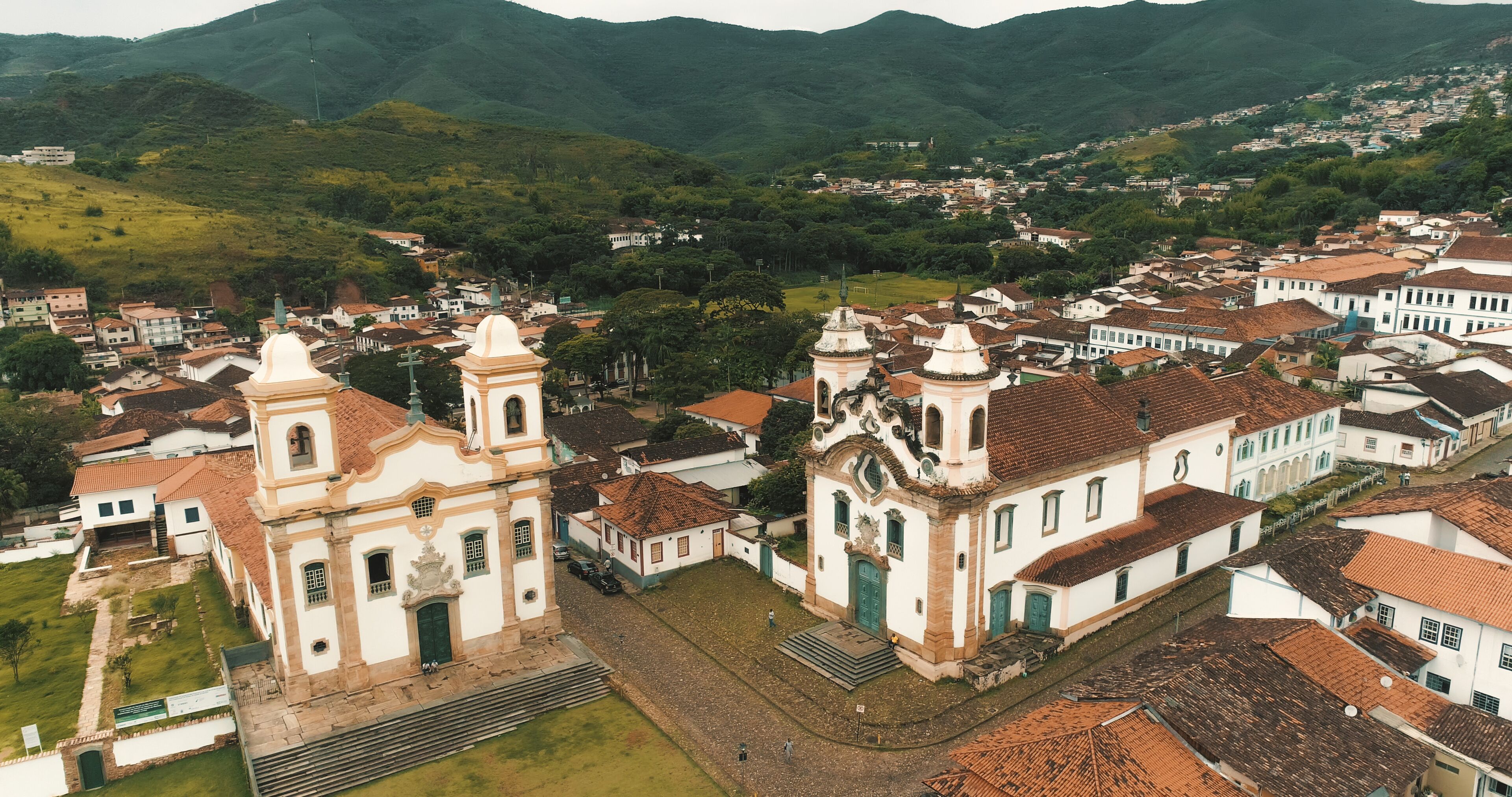 An aerial view of a town with historic buildings, including a church with two bell towers, surrounded by lush green hills.