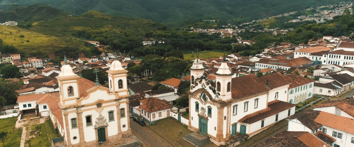 An aerial view of a town with historic buildings, including a church with two bell towers, surrounded by lush green hills.