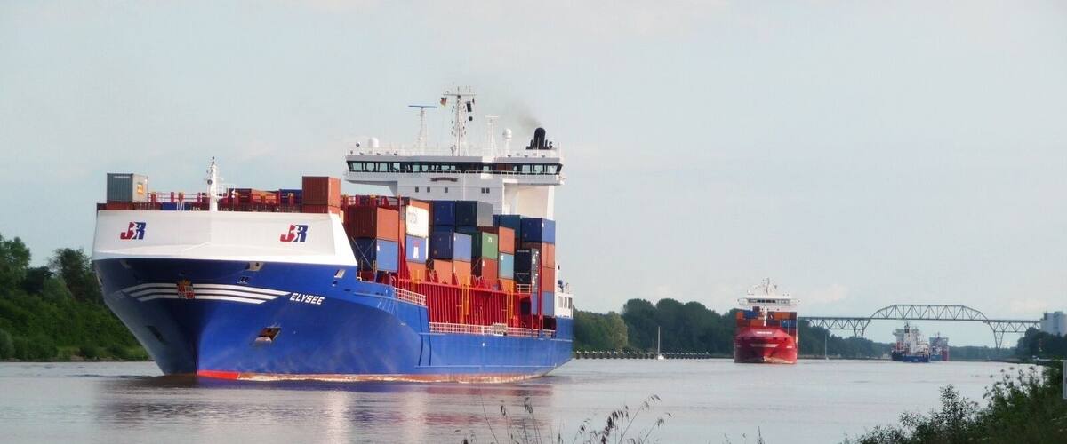 A large cargo ship with multiple containers, a smaller tugboat, and a bridge in the background.