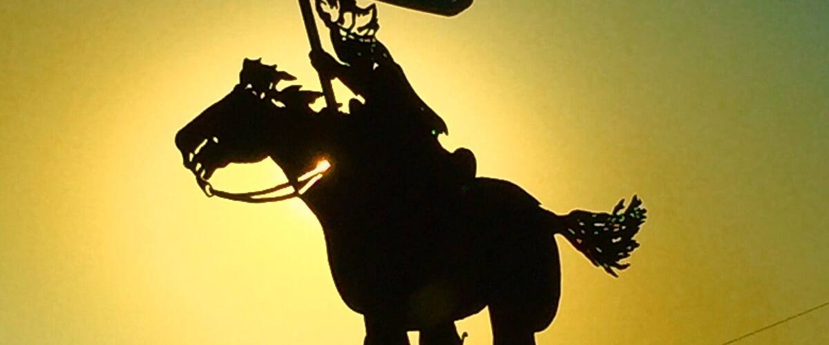A silhouette of a horse statue with a flag, set against a sunset.