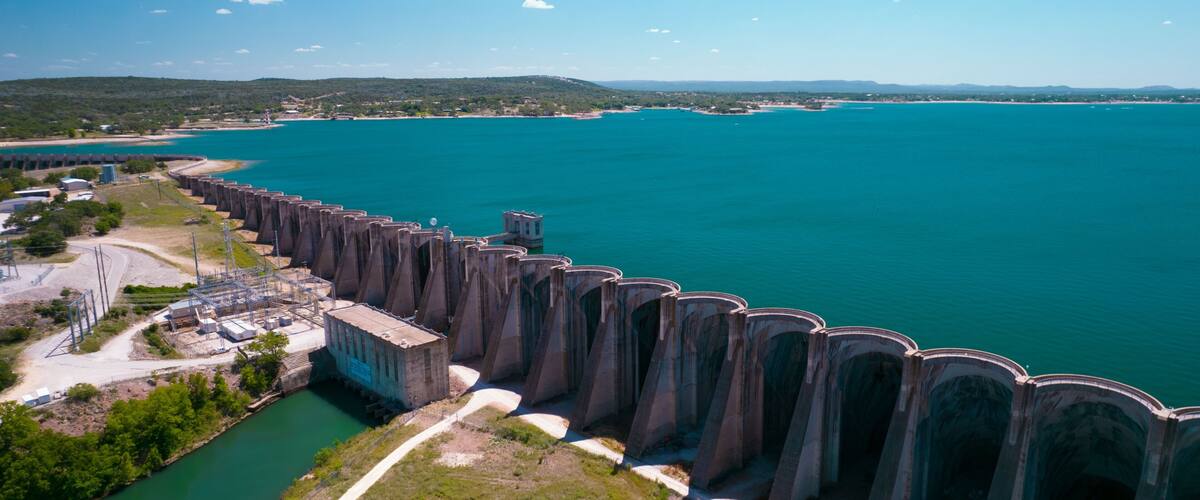 A large dam with multiple spillways, a river, and a coastal area with buildings and greenery.