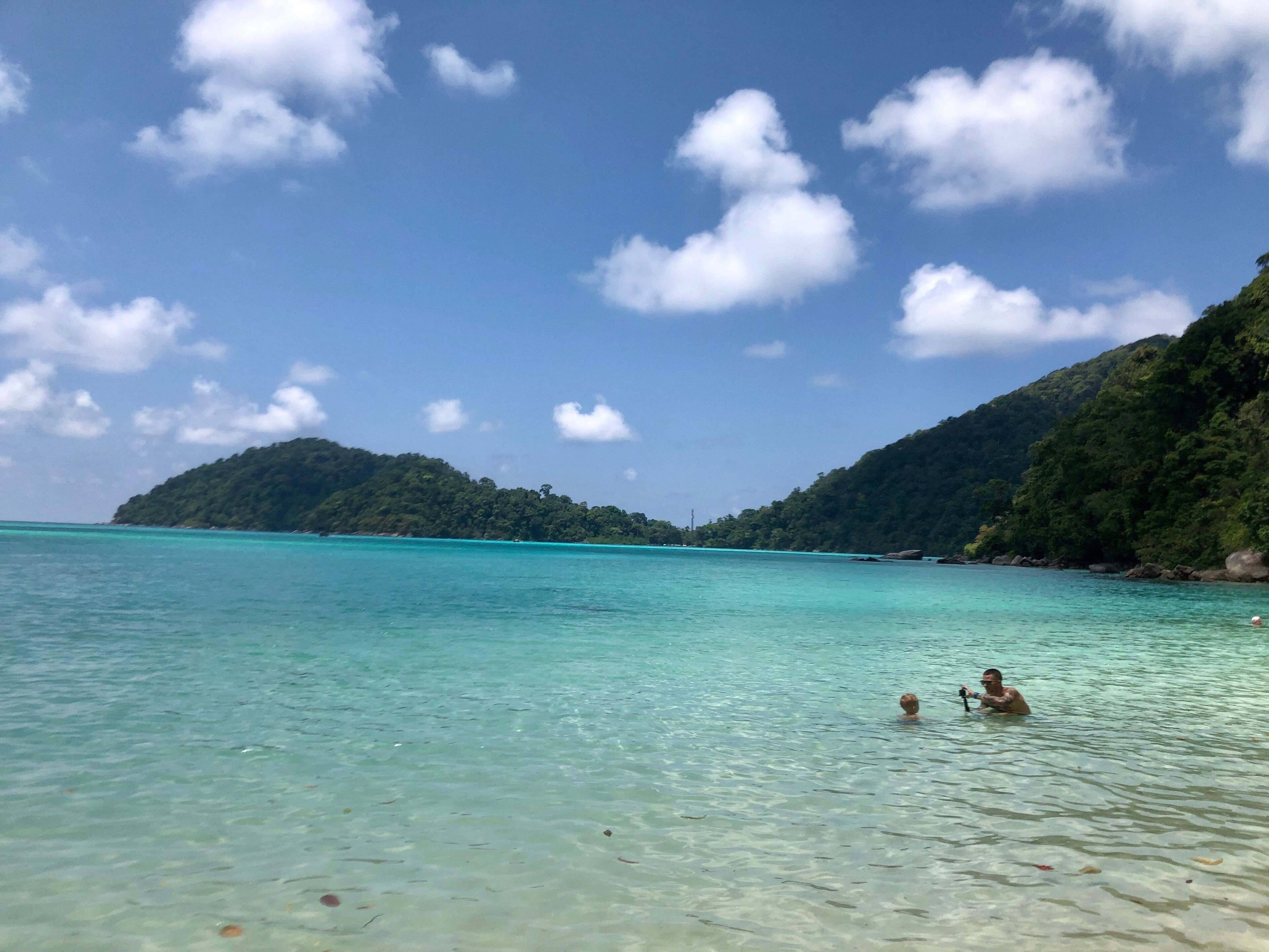 A clear turquoise sea with a person swimming and another person taking a photo. There are green hills in the background.